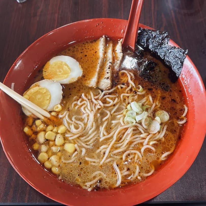 A vibrant red bowl of ramen with noodles, pork, corn, green onions, nori, and two soft-boiled eggs.