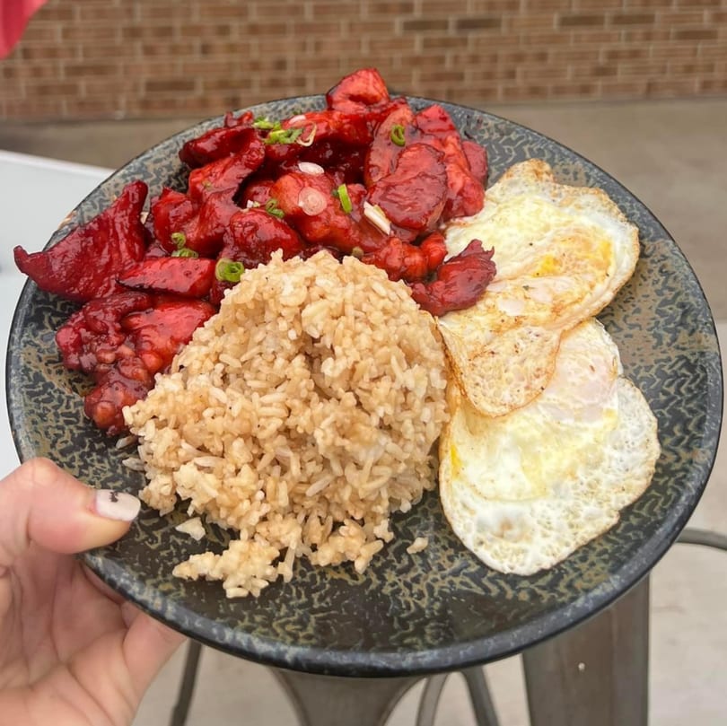 A plate of red meat, fried rice, and two fried eggs, held by a hand.