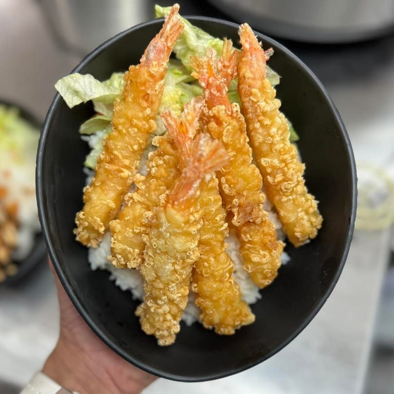 A hand holds a black bowl of golden tempura shrimp over white rice and lettuce.